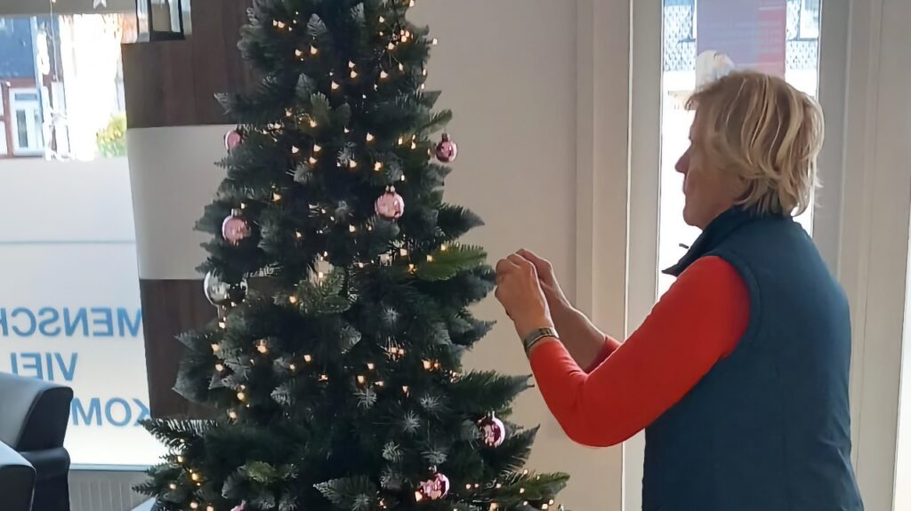 Eine Frau in einem roten Oberteil und einer dunklen Weste schmückt einen künstlichen Weihnachtsbaum mit Lichterketten und rosa Weihnachtskugeln. Der Baum steht in einem hellen Eingangsbereich.