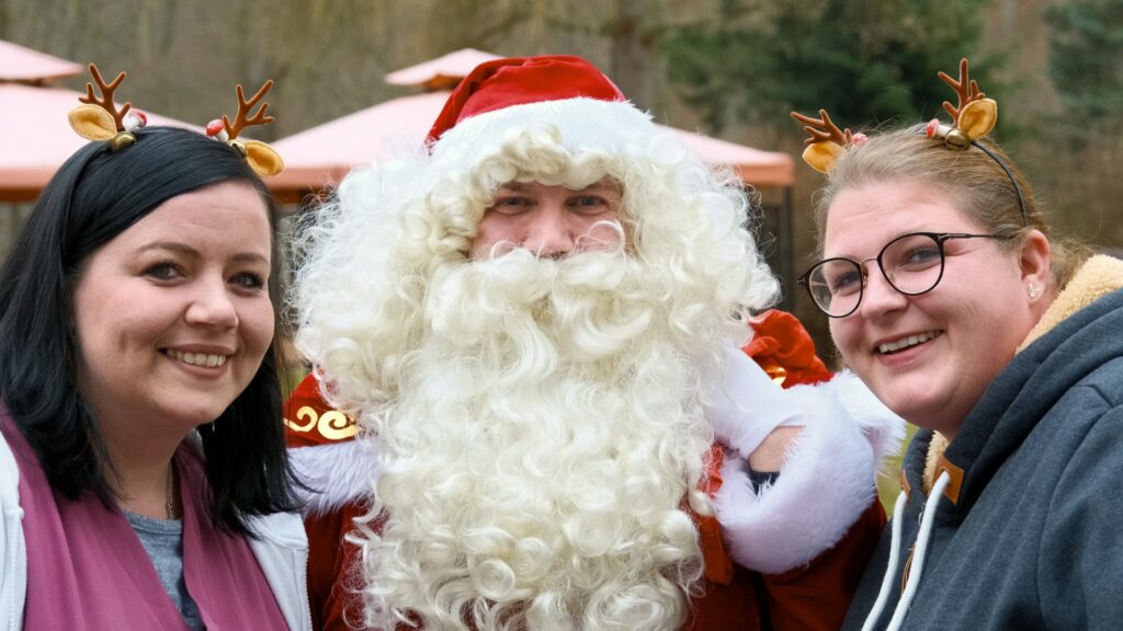 Zwei lächelnde Frauen, die Rentier-Haarreifen tragen, posieren im Freien gemeinsam mit dem Weihnachtsmann für ein Foto.