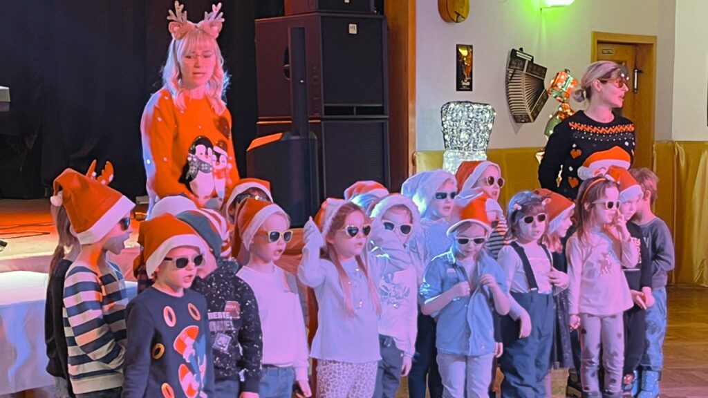 Eine Gruppe von Kindern der Kita „Wirbelwind“ steht auf einer Bühne und singt. Sie tragen rote Weihnachtsmützen und Sonnenbrillen. Zwei Erzieherinnen begleiten den Auftritt im Hintergrund.