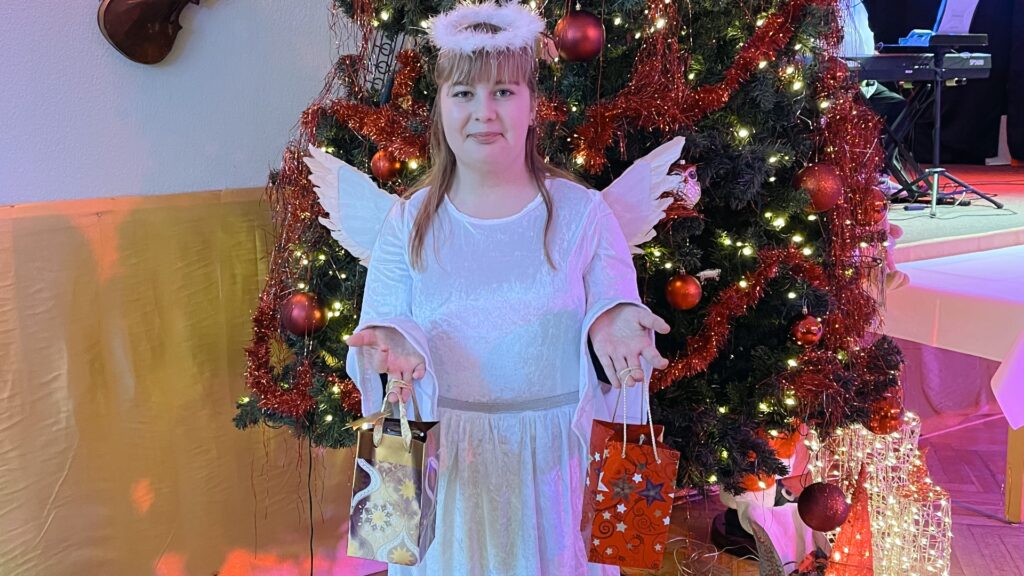 Ein Mädchen, verkleidet als Engel mit weißen Flügeln, Heiligenschein und weißem Gewand, steht vor einem geschmückten Weihnachtsbaum und hält zwei Geschenktüten in den Händen.
