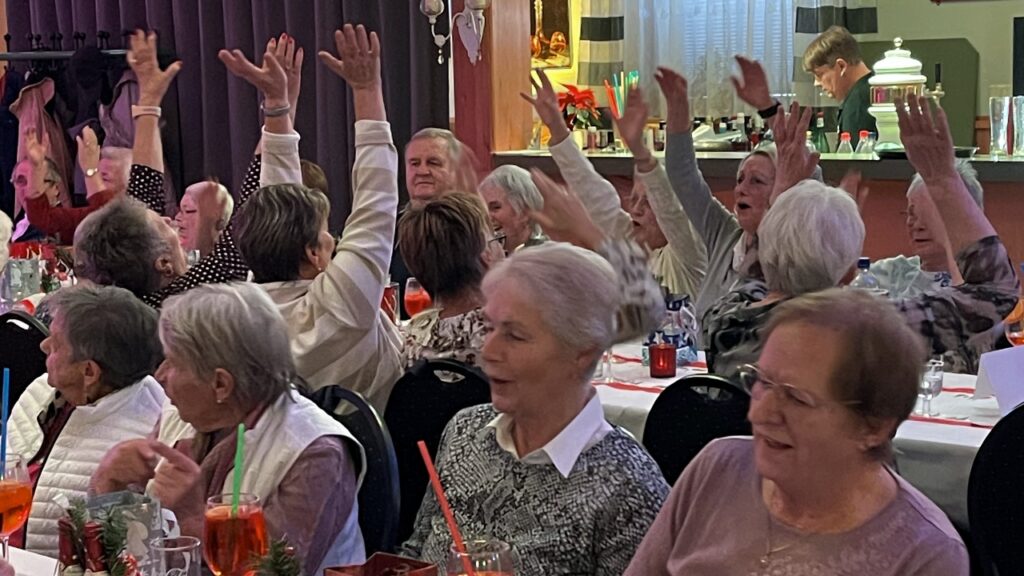 Zahlreiche Seniorinnen und Senioren sitzen an festlich gedeckten Tischen im Saal des „Sachswerfer Handwagens“. Sie haben ihre Hände freudig in die Luft gehoben und machen bei der Unterhaltung mit.