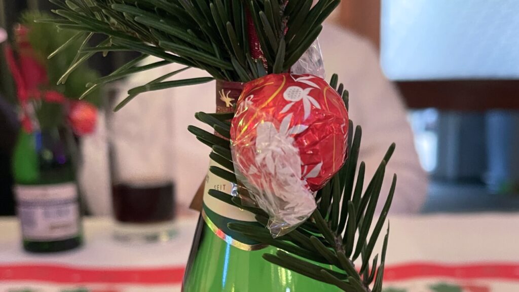 Nahaufnahme einer grünen Getränkeflasche auf einem Tisch, die liebevoll mit einem kleinen Tannenzweig und einer rot verpackten Schokoladenkugel dekoriert ist.