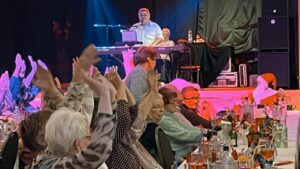 Der Musiker Achim Großmann sitzt auf der beleuchteten Bühne an einem Keyboard und singt. Im Vordergrund klatschen die Senioren begeistert im Takt mit.