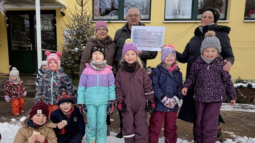 Zwei Frauen, ein Mann und mehrere Kinder vor einem Kindergartengebäude, ein Scheck wird in die Kamera gehalten.