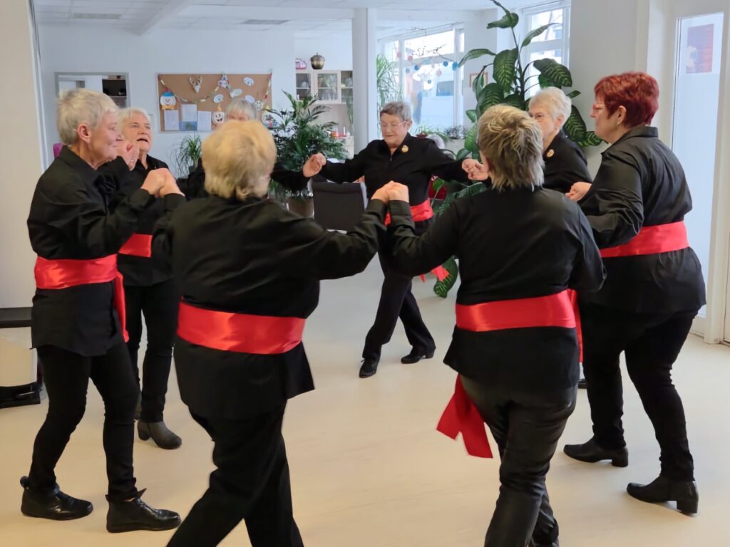 Eine Gruppe von Seniorinnen tanzt im Kreis in einem hellen Raum. Sie tragen einheitlich schwarze Blusen und Hosen sowie leuchtend rote Schärpen um die Hüfte.