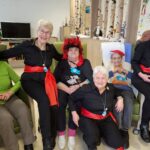 Fünf gut gelaunte Frauen bei der Faschingsfeier. Zwei Frauen der Tanzgruppe stehen außen, in der Mitte sitzen drei Seniorinnen mit lustigen Kopfbedeckungen wie Teufelshörnern und roten Perücken.