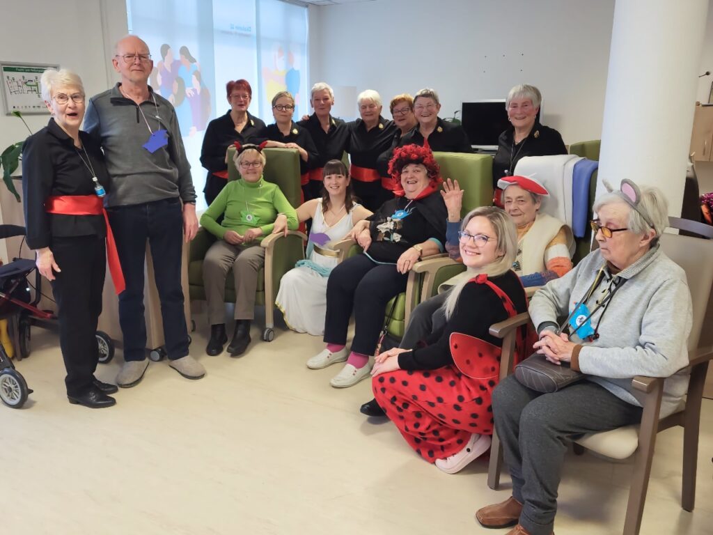 Gruppenfoto einer Faschingsfeier im Pflegeheim: Bewohner und Mitarbeiter sind bunt verkleidet, unter anderem als Marienkäfer, Teufel oder Maus. Im Hintergrund steht eine Tanzgruppe in schwarzer Kleidung.