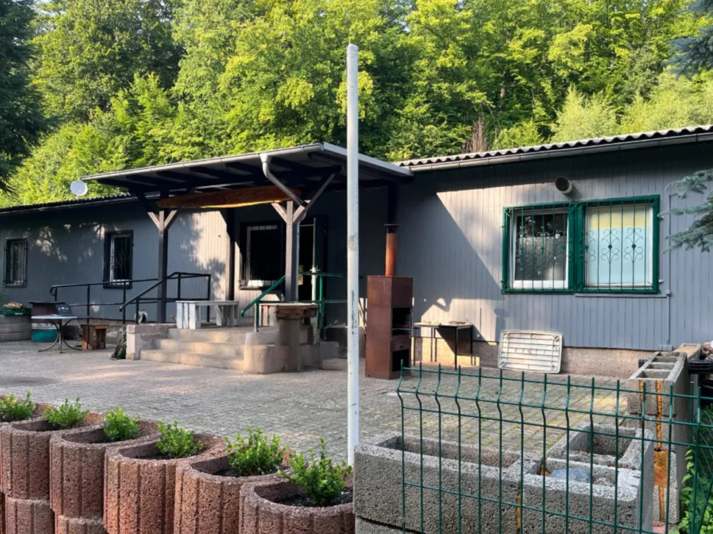 Außenansicht des Vereinsheims „MADHOUSE“ in Ilfeld. Es handelt sich um ein einstöckiges, grau verkleidetes Gebäude mit einem flachen Vordach über dem Eingangsbereich. Vor dem Haus befindet sich ein gepflasterter Vorplatz mit einem Außenkamin und Pflanzkübeln im Vordergrund. Das Gebäude liegt idyllisch am Waldrand mit dichten grünen Bäumen im Hintergrund.