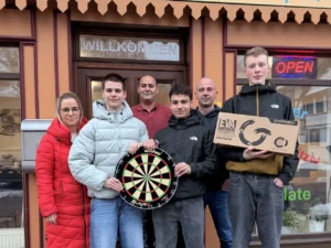 Gruppenfoto von sechs Personen vor einem Gebäude mit einem 'WILLKOMMEN'-Schild über der Tür. Im Vordergrund präsentieren drei Jugendliche stolz neues Darts-Zubehör: eine Dartscheibe und einen originalverpackten Schutzring. Flankiert werden sie von zwei Männern im Hintergrund und einer Frau in einem auffälligen roten Wintermantel auf der linken Seite.