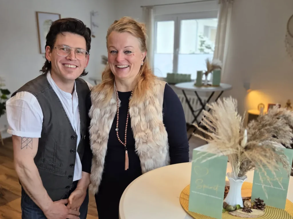 Ein lächelndes Paar steht in einem hellen Raum. Der Mann links trägt eine dunkle Weste über einem weißen Hemd und eine Brille. Die Frau rechts trägt ein dunkles Oberteil mit einer hellen Kunstfellweste. Im Vordergrund auf einem Tisch steht eine grüne Grußkarte mit der Aufschrift 'Spirit'.