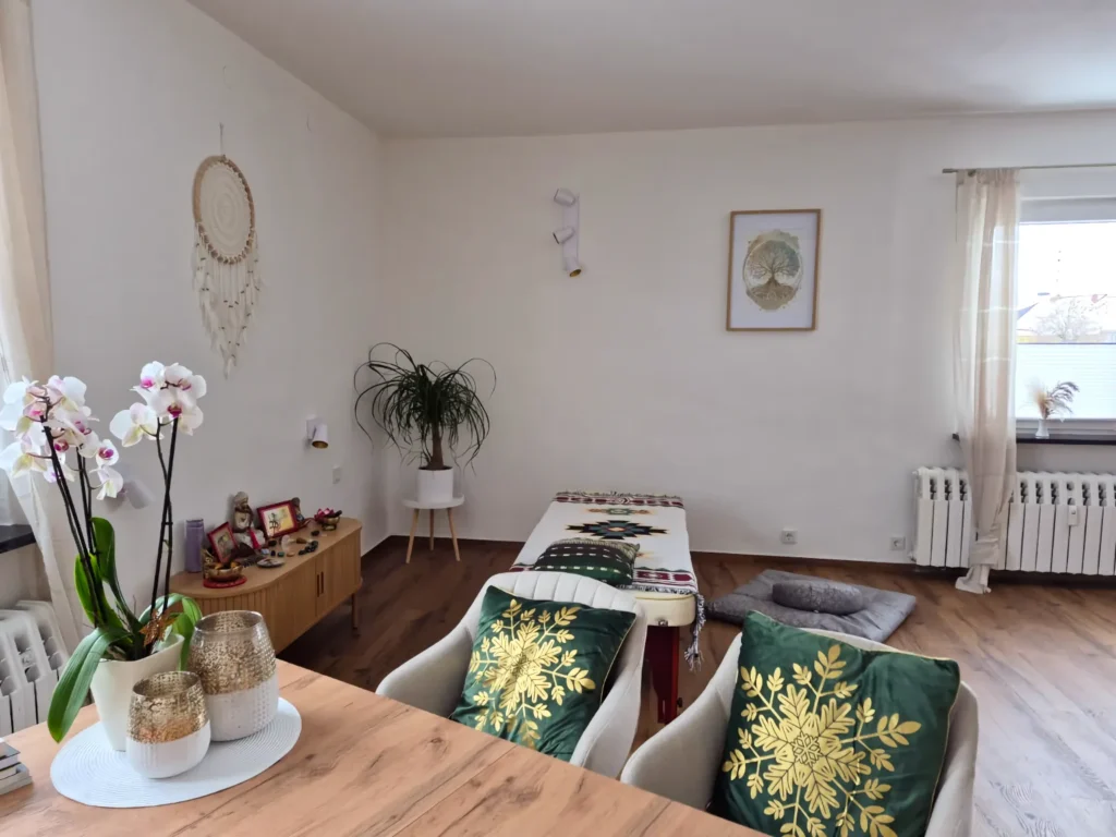 Ein heller Raum mit Holzboden. Im Vordergrund stehen zwei graue Sessel mit grünen Kissen an einem Holztisch, auf dem Orchideen stehen. Hinten im Raum befindet sich eine Behandlungsliege. An der weißen Wand hängt ein Traumfänger.