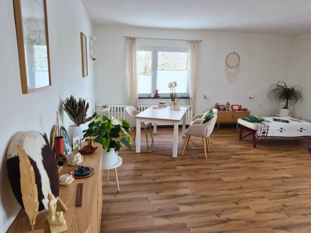 Ein weitläufiger, gemütlich eingerichteter Raum mit Holzboden. In der Mitte steht ein heller Esstisch mit Stühlen. Im Hintergrund rechts befindet sich eine Behandlungsliege. Auf einem Sideboard im Vordergrund liegen verschiedene Deko-Elemente.