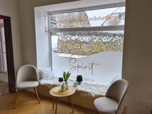 Ein Fensterplatz mit zwei grauen Sesseln und einem kleinen Tisch. Die untere H&auml;lfte der Fensterscheibe ist milchig foliert und tr&auml;gt den goldenen Schriftzug 'Spirit - Zentrum f&uuml;r ganzheitliche Heilbehandlungen'. Auf der Fensterbank liegen flauschige wei&szlig;e Kissen.
