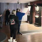 Innenansicht des Vereinsheims während eines Soft-Dart-Spiels. Mehrere Personen stehen in einem Raum mit dunklem Boden vor modernen Soft-Dart-Automaten, die an einer Wand in Backsteinoptik aufgestellt sind. Ein Spieler im blauen T-Shirt zielt gerade auf eine der leuchtenden Dartscheiben. Über den Geräten hängt eine kleine Discokugel an der Decke.
