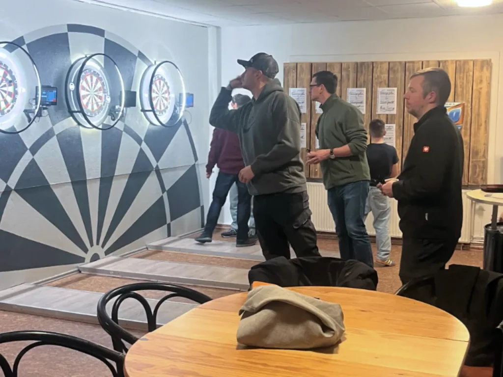 Drei Männer beim Steel-Dart-Training im Innenraum des Vereins. An einer weißen Wand, die mit einer großen Zielscheiben-Grafik bemalt ist, hängen mehrere professionelle Steel-Dart-Boards mit LED-Beleuchtungsringen. Im Vordergrund steht ein Holztisch; die Stimmung ist konzentriert und sportlich.