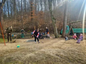 Mehrere Erwachsene und Kinder bei einem Arbeitseinsatz auf einem Waldspielplatz. Im Vordergrund harken eine Frau und ein Mann den Sandboden, im Hintergrund spielen Kinder auf einer Schaukel und ein kleiner gr&uuml;ner Lastwagen der Gemeinde steht am Waldrand. Die Sonne scheint hell durch die hohen, noch kahlen B&auml;ume im Hintergrund.