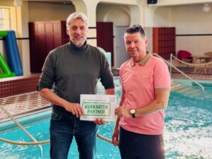 B&uuml;rgermeister Stephan Klante (links) und Badehaus-Gesch&auml;ftsf&uuml;hrer Jens Eisenschmidt (rechts) stehen im Hallenbad des Badehaus Nordhausen und halten gemeinsam das gr&uuml;n-wei&szlig;e Kurkarten-Partner-Schild der Gemeinde Harztor in die Kamera.
