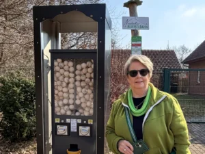 Eine Frau in einer gr&uuml;nen Jacke steht l&auml;chelnd neben einem schwarzen Murmelautomaten im Freien. Der Automat ist mit vielen hellen Holzkugeln gef&uuml;llt und befindet sich unter einer neu installierten, anthrazitfarbenen Metall&uuml;berdachung auf einem gemauerten Sockel. Im Hintergrund ist ein Schild mit der Aufschrift &bdquo;Neust&auml;dter Murmelbahn&ldquo; zu sehen.