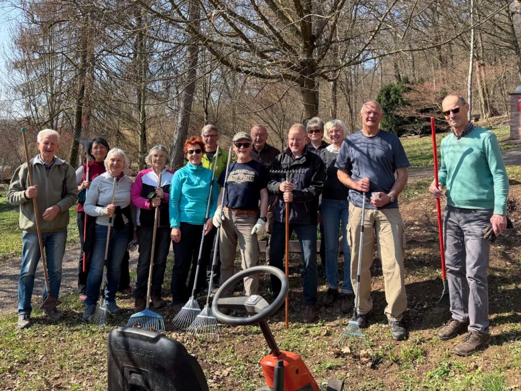 Gruppenfoto von rund 15 Helferinnen und Helfern mit Rechen und Gartengeräten im Ahornpark Ilfeld. Die Teilnehmer stehen in einer Reihe auf der Parkwiese. Im Vordergrund ist das Lenkrad eines Husqvarna-Rasentraktors zu sehen.