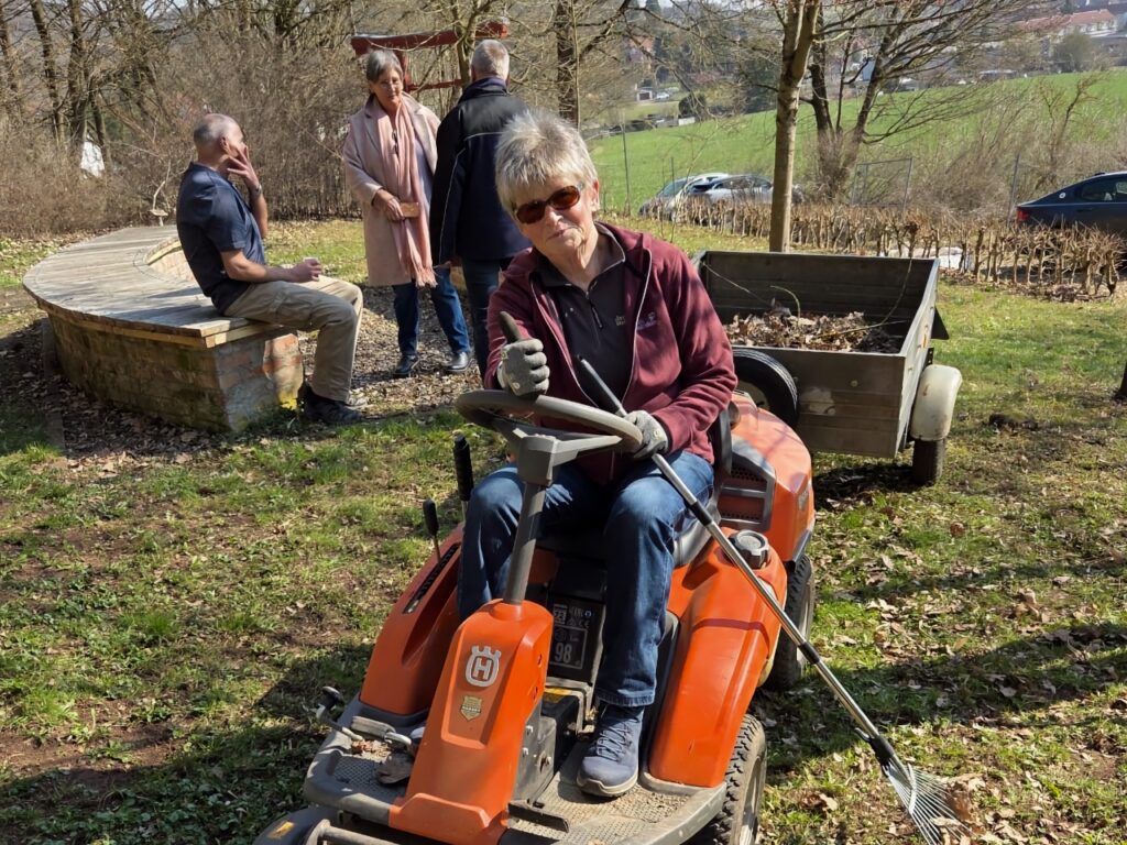 Eine Helferin sitzt auf einem orangefarbenen Husqvarna-Rasentraktor mit Anhänger im Ahornpark Ilfeld. Im Hintergrund unterhalten sich weitere Helfer an einem runden Sitzpodest aus Holz und Stein.