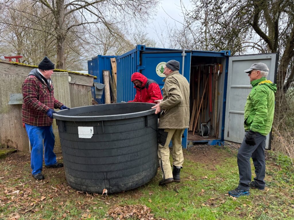 Vier Männer bewegen gemeinsam ein großes schwarzes Wasserfass vor einem blauen Container mit dem Logo des Ahornparks. Der Container dient als Geräteschuppen mit Gartengeräten.