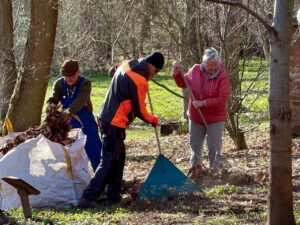 Drei Helfer beim Frühjahrsputz im Ahornpark Ilfeld. Zwei Männer sammeln Herbstlaub in einen großen weißen Sack, eine Frau in roter Jacke harkt mit einem Laubrechen. Im Hintergrund kahle Laubbäume und erstes Grün auf der Wiese.