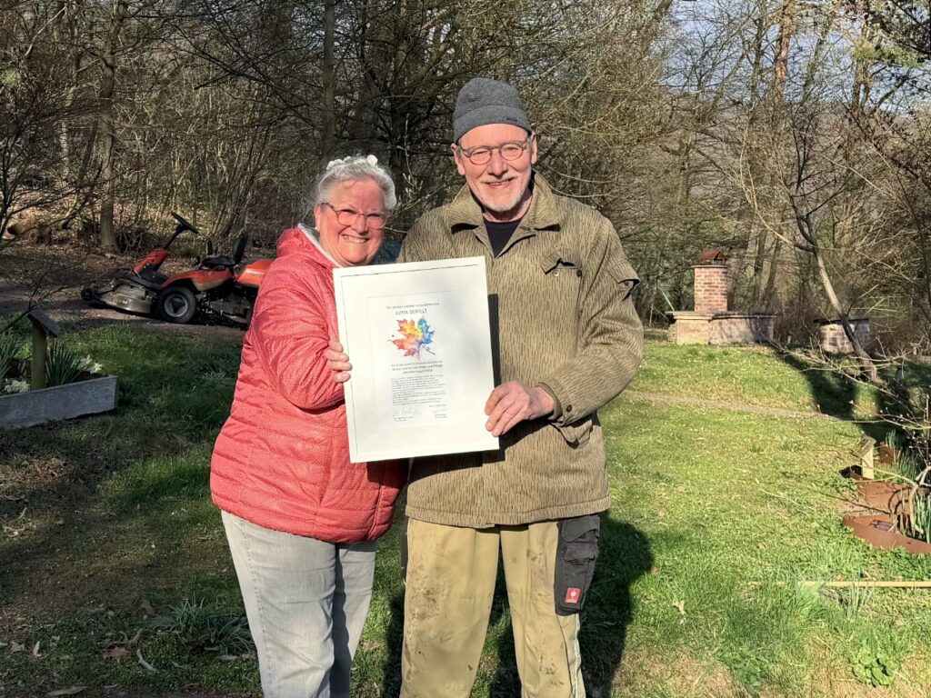 Eine Frau in roter Jacke und ein Mann in olivfarbener Jacke halten gemeinsam eine gerahmte Urkunde mit buntem Ahornblatt-Motiv in die Kamera. Im Hintergrund der Ahornpark mit einem Aufsitzmäher und erstem Frühlingsgrün.