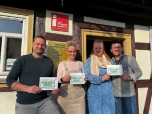 Vier Personen stehen l&auml;chelnd vor dem Eingang der Tourismus-Information Neustadt und halten jeweils ein gr&uuml;nes Schild mit der Aufschrift &bdquo;Kurkarten-Partner" in die Kamera. Von links: Roland Gerlach, Alexandra G&uuml;nther, Ariane Bruchmann und Sascha Niedenf&uuml;hr.
