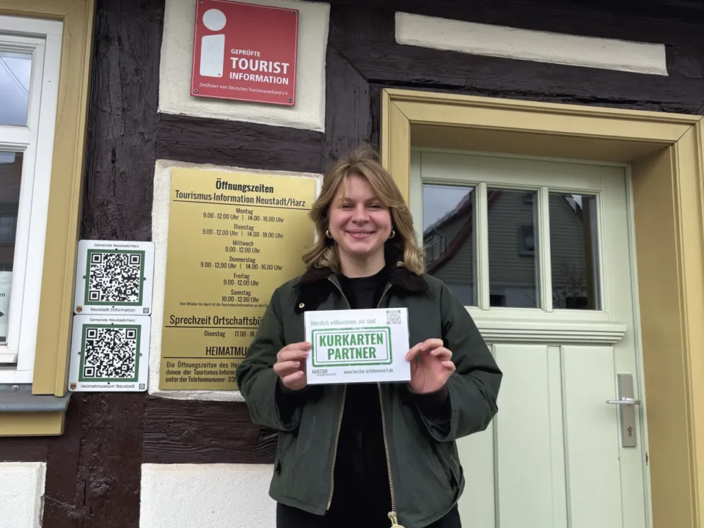 Eine junge Frau mit hellblondem Haar und grüner Jacke steht lächelnd vor dem Eingang der Tourismus-Information Neustadt/Harz und hält eine weiße Karte mit grünem Schriftzug „Kurkarten-Partner" in die Kamera. Im Hintergrund sind das Öffnungszeiten-Schild der Tourismus-Information sowie das Qualitätssiegel „Geprüfte Tourist Information" des Deutschen Tourismusverbandes sichtbar. Abgebildet: Veronika Töpfer, Prokuristin und Standortleiterin der HEX Erlebniswelt Rothesütte.