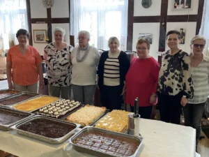 Sieben Frauen stehen nebeneinander hinter einem langen Tisch, auf dem mehrere gro&szlig;e Backbleche mit verschiedenen selbst gebackenen Kuchen angerichtet sind. Im Hintergrund ist ein Fachwerkraum mit wei&szlig;en Gardinen zu sehen.