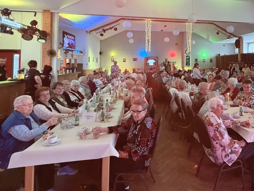 Ein großer, festlich geschmückter Veranstaltungssaal mit nahezu vollständig besetzten Tischreihen. Die Gäste sitzen an weißen gedeckten Tischen mit Kerzen. Bunte Lichteffekte und weiße Lampions an der Decke sorgen für festliche Atmosphäre.