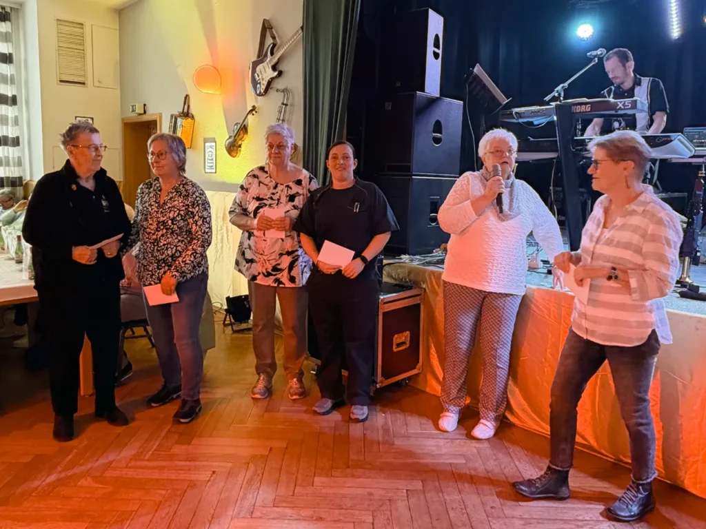 Sechs Personen stehen bei einer Dankes- und Ehrungsszene nahe der Bühne. Eine ältere Frau spricht ins Mikrofon, während die anderen Personen Umschläge oder Gutscheine in der Hand halten. Im Hintergrund spielt ein Keyboarder auf der Bühne.