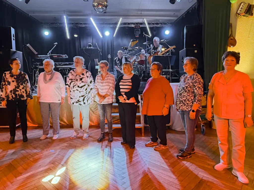 Acht Frauen stehen in einer Reihe auf einem Parkettboden vor einer beleuchteten Bühne, auf der eine Band spielt. Der Saal ist in warmes Bühnenlicht getaucht; im Hintergrund ist eine Discokugel sichtbar.