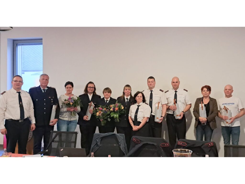 Gruppenfoto vom Feuerwehrverein Niedersachswerfen: Elf Personen stehen nebeneinander in einem Versammlungsraum, mehrere in Feuerwehruniform mit weißem Hemd und dunkler Krawatte, andere in Alltagskleidung. Einige halten Blumensträuße und kleine Geschenkpäckchen in den Händen. Im Vordergrund ist ein Sitzungstisch mit abgelegten Uniformjacken zu sehen – eine freundliche Stimmung nach den Neuwahlen des Vorstandes.