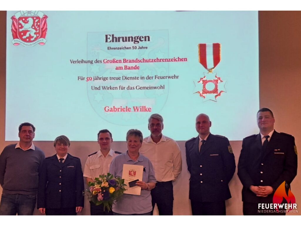 Innenaufnahme im Feuerwehr-Versammlungsraum: Gabriele Wilke steht mittig und hält eine Urkunde sowie einen Blumenstrauß in den Händen. Links neben ihr steht ein Feuerwehrmann in dunkelblauer Dienstuniform mit weißem Hemd, der ihr gratuliert; rechts ein weiterer uniformierter Kamerad. Im Hintergrund ist das Feuerwehrwappen an der Wand zu erkennen – ein feierlicher Moment zur Verleihung des Großen Brandschutzehrenzeichens für 50 Jahre treue Dienste.