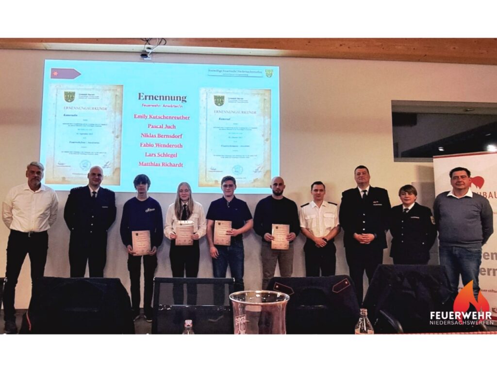 Gruppenfoto im Feuerwehr-Versammlungsraum: Die neu ernannten Feuerwehr-Anwärterinnen und -Anwärter stehen nebeneinander, jeweils mit Ernennungsurkunden in der Hand. Neben ihnen steht die Wehrführung in dunkelblauer Dienstuniform. Zu sehen sind Emily Kutschenreuther, Pascal Juch, Niklas Bernsdorf, Fabio Wenderoth, Lars Schlegel und Mathias Richardt – sichtbar stolz in ihrem ersten offiziellen Moment als Teil der Einsatzabteilung.