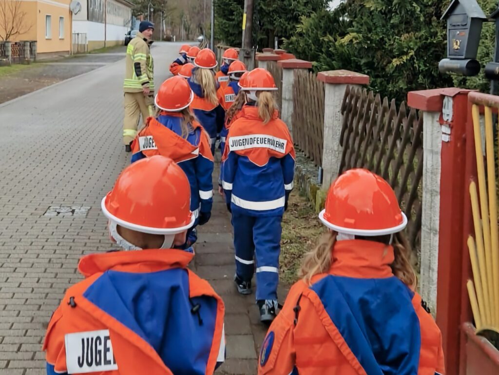 Mehrere Kinder der Jugendfeuerwehr Niedersachswerfen in blau-oranger Einsatzkleidung mit orangefarbenen Helmen laufen in einer Reihe über einen Gehweg einer Dorfstraße. An der Spitze der Gruppe geht ein Feuerwehrmann in gelber Einsatzjacke. Im Hintergrund sind Wohnhäuser und Gärten zu sehen.