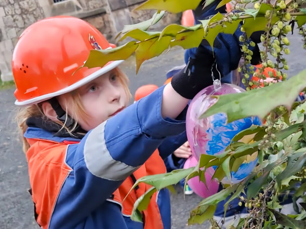 Nahaufnahme eines Kindes der Jugendfeuerwehr in blau-oranger Einsatzjacke und orangefarbenem Helm, das konzentriert ein bunt bemaltes Osterei aus Glas an einem Zweig mit grünen Mahonienblättern befestigt. Im Hintergrund ist das alte Mauerwerk der Kirche Niedersachswerfen zu erkennen.