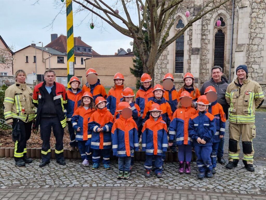 Gruppenfoto vor der Kirche Niedersachswerfen: Rund fünfzehn Kinder der Jugendfeuerwehr in blau-orangen Uniformen mit Helmen stehen gemeinsam mit vier Betreuerinnen und Betreuern in Einsatzkleidung neben dem mit bunten Ostereiern geschmückten Baum. Im Hintergrund das Kirchengebäude und ein festlich gestalteter Osterbrunnen-Platz.