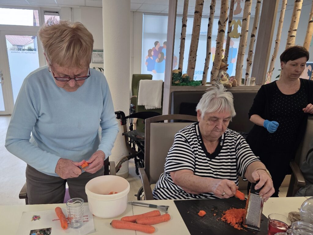 Helle, freundliche Innenaufnahme aus der Tagespflege St. Johannis in Niedersachswerfen: Links steht eine Seniorin mit hellblauem Pullover und Brille und schält konzentriert eine Möhre über einer weißen Schüssel. Rechts reibt eine zweite Seniorin im schwarz-weiß gestreiften Oberteil eine Möhre auf einer kleinen Metallreibe. Auf dem Tisch liegen weitere Möhren, eine Gabel und ein Schneidebrett. Im Hintergrund sind ein Pflegerollator, dekorative Birkenstämme und österliche Fensterbilder zu sehen – rechts betreut eine Pflegekraft mit blauen Einweghandschuhen die Szene.