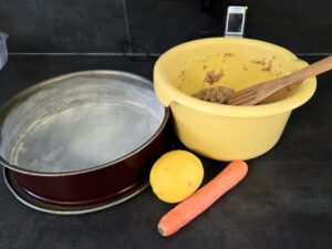 Stillleben auf einer dunklen K&uuml;chenarbeitsplatte: Links steht eine runde Springform, bereit zum Bef&uuml;llen. Rechts daneben eine gelbe R&uuml;hrsch&uuml;ssel mit dem vorbereiteten Karottenkuchen-Teig und einem Holzl&ouml;ffel. Davor liegen eine gelbe Zitrone und eine frische M&ouml;hre &ndash; die charakteristischen Zutaten f&uuml;r einen saftigen Karottenkuchen warten auf den n&auml;chsten Arbeitsschritt.