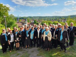 Gruppenfoto des Gemischten Chores Ellrich: Rund 45 Sängerinnen und Sänger in festlicher dunkler Kleidung mit türkisfarbenen Schals stehen auf einer Anhöhe. Im Hintergrund ist ein Ortspanorama mit Kirchturm, Fachwerkhäusern und grünen Hügeln zu sehen. Sonniger Tag mit blauem Himmel und vereinzelten Wolken.