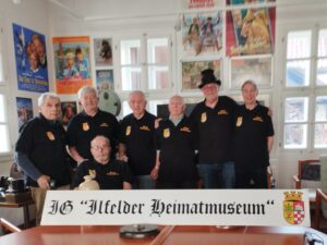 Sieben aktive Mitglieder der IG Ilfelder Heimatmuseum stehen und sitzen in einem Ausstellungsraum des Museums. Alle tragen schwarze Polo-Shirts mit dem gelb-roten Wappen und der Aufschrift &bdquo;Ilfelder Heimatmuseum". Im Vordergrund halten sie ein wei&szlig;es Schild mit der Aufschrift &bdquo;IG Ilfelder Heimatmuseum" in altdeutscher Schrift, dazu das Wappen von Ilfeld. An den W&auml;nden hinter ihnen h&auml;ngen historische Filmplakate. Von links nach rechts: Gerhardt Hosbach, Achim Fiebig, Karl-Heinz Bruchmann, Fred Minerup, Walter Albrecht, Klaus Liebenrodt, Manfred B&ouml;ttcher (sitzend). Dieter Z&ouml;rle ist auf dem Foto nicht abgebildet.