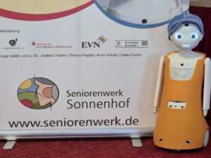 Empathie-Roboter Navel steht vor einem Roll-up-Banner des Seniorenwerks Sonnenhof. Er hat ein wei&szlig;es Gesicht mit gro&szlig;en, blau leuchtenden Augen und einen freundlichen Mund. Auf dem Kopf tr&auml;gt er eine gestrickte blaue M&uuml;tze mit aufgesticktem Buchstaben N. Sein K&ouml;rper ist orange. Auf dem Banner im Hintergrund sind Logos der Unterst&uuml;tzer zu sehen, darunter EVN Nordhausen, Stiftung der Kreissparkasse Nordhausen und Autohaus Gerecke.