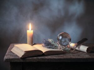Ein dunkler Holztisch mit einem aufgeschlagenen, in Leder gebundenen Buch mit vergilbten Seiten. Daneben brennt eine lange Bienenwachskerze. Vor dem Buch liegen ein kleines B&uuml;ndel getrockneter Kr&auml;uter, eine gl&auml;nzende Kristallkugel und eine Schreibfeder auf einer Pergamentrolle. Warmes Kerzenlicht und weiche Schatten erzeugen eine geheimnisvolle Stimmung. KI.