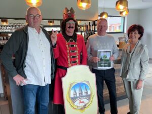 Vier Personen stehen nebeneinander im modernen Restaurantbereich des Golfparks Neustadt. Die zweite Person von links tr&auml;gt ein rotes Karnevalskost&uuml;m mit Krone und h&auml;lt ein dekoratives Wappenschild mit dem Ortswappen von Neustadt. Die dritte Person h&auml;lt eine Urkunde mit der Aufschrift: &bdquo;Der Ortschaftsrat Neustadt sponsert ein Baum f&uuml;r den Golfpark Neustadt &ndash; Wert: 500 &euro;". Im Hintergrund sind Weinregale und H&auml;ngeleuchten zu sehen.