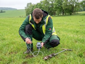 Symbolfoto Bodenluftmessung: Ein Techniker in Arbeitskleidung entnimmt mit einer Bodensonde eine Bodenluftprobe. Er tr&auml;gt Handschuhe und eine Warnweste. Die Sonde steckt in einer gr&uuml;nen Wiese. Im Hintergrund sind B&auml;ume zu sehen. Symbolfoto f&uuml;r die Radonmessungen in Harztor. KI.