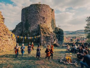 Blick auf den rund 19 Meter hohen Bergfried der Ruine Ebersburg aus r&ouml;tlich-violettem Porphyrit. Der massive Turmstumpf mit seinen knapp f&uuml;nf Meter dicken Mauern ragt &uuml;ber das Krebsbachtal bei Herrmannsacker. Im Vordergrund spielen Kinder in mittelalterlichen Kost&uuml;men auf der Wiese der Burganlage. Bunte Wimpel sind zwischen den erhaltenen Mauerresten gespannt.