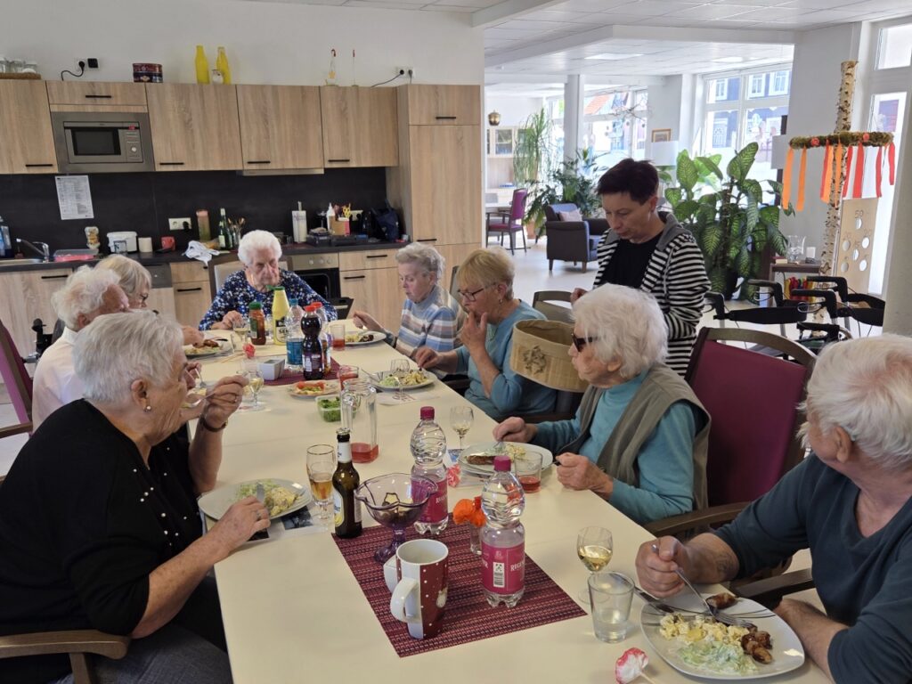 Gruppe älterer Menschen beim gemeinsamen Mittagessen an einem langen Tisch, betreut von einer Pflegekraft.