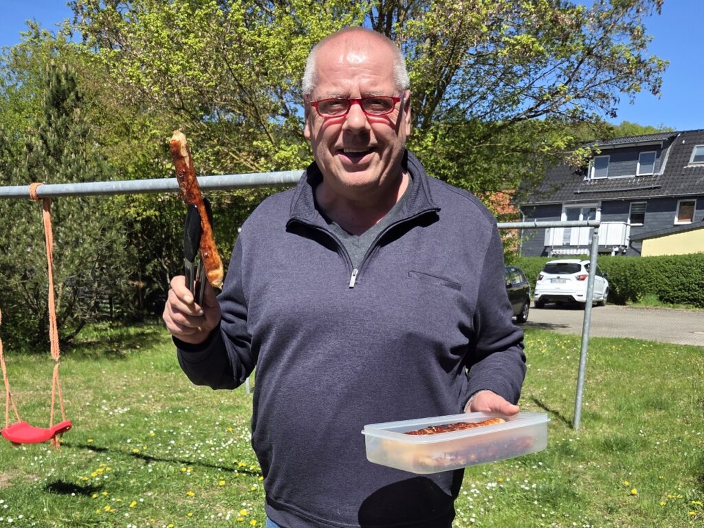 Mann im Freien hält lachend eine Bratwurst mit einer Grillzange und eine Vorratsdose mit mariniertem Fleisch.